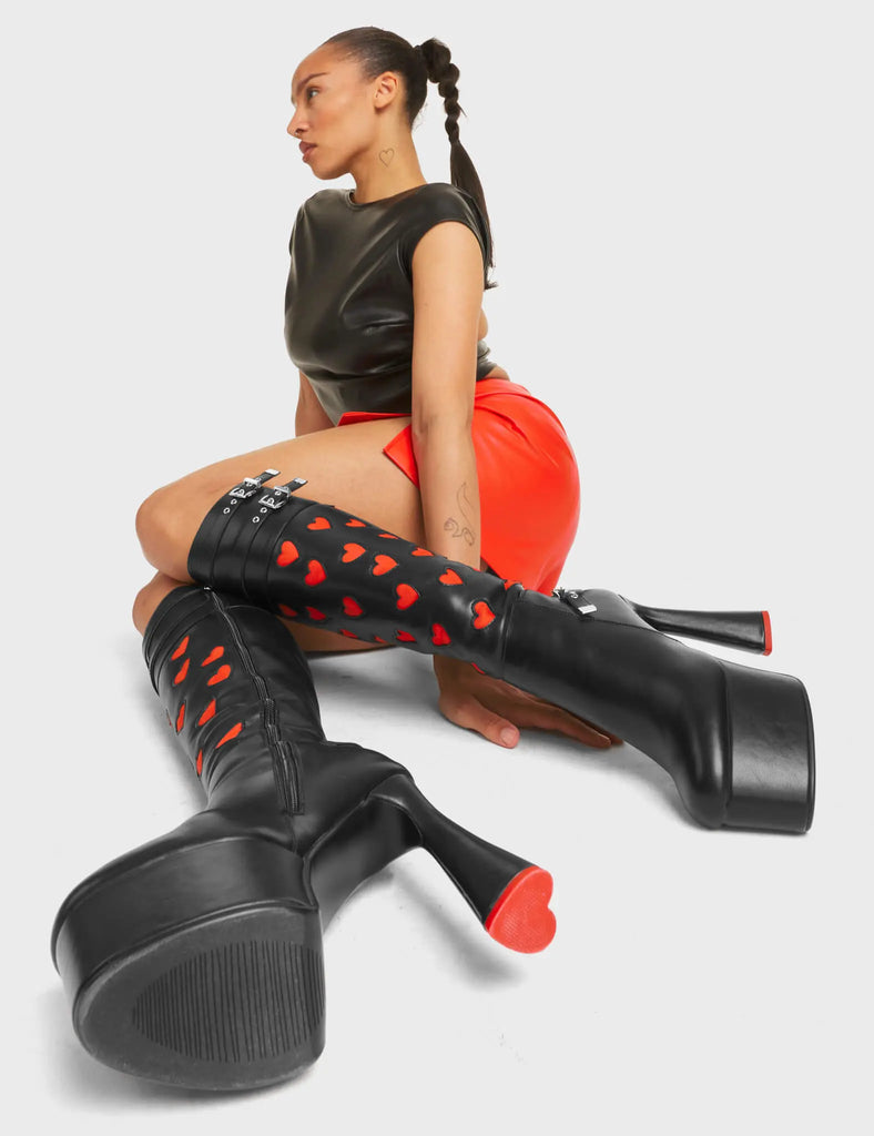 Talk Platform Knee High Boots in Black Stretch faux leather. These platform boots feature a panel of red heart-patterned material, with adjustable straps at the top and on the ankle of the boot. The straps have silver hardware including square-shaped buckles, and the platform sole features a red heart-shaped heel. Made with eco-friendly materials and 100% cruelty-free!
