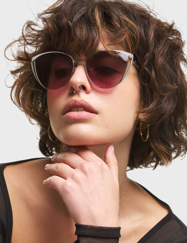 Rosa Cateye Sunglasses. These Cateye sunglasses feature a Silver frame with Silver Glitter eyebrows, and a dark Pink tinted lens.
