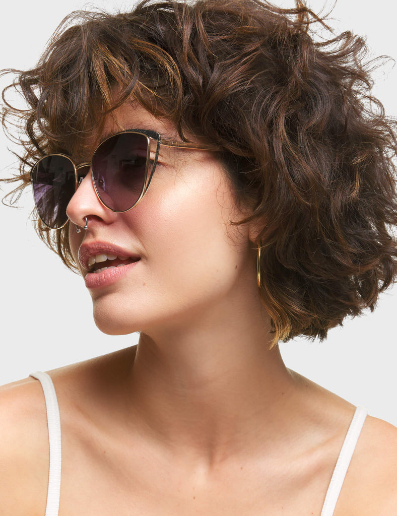 Rosa Cateye Sunglasses. These Cateye sunglasses feature a Gold frame with Black Glitter eyebrows, and a dark Purple tinted lens.