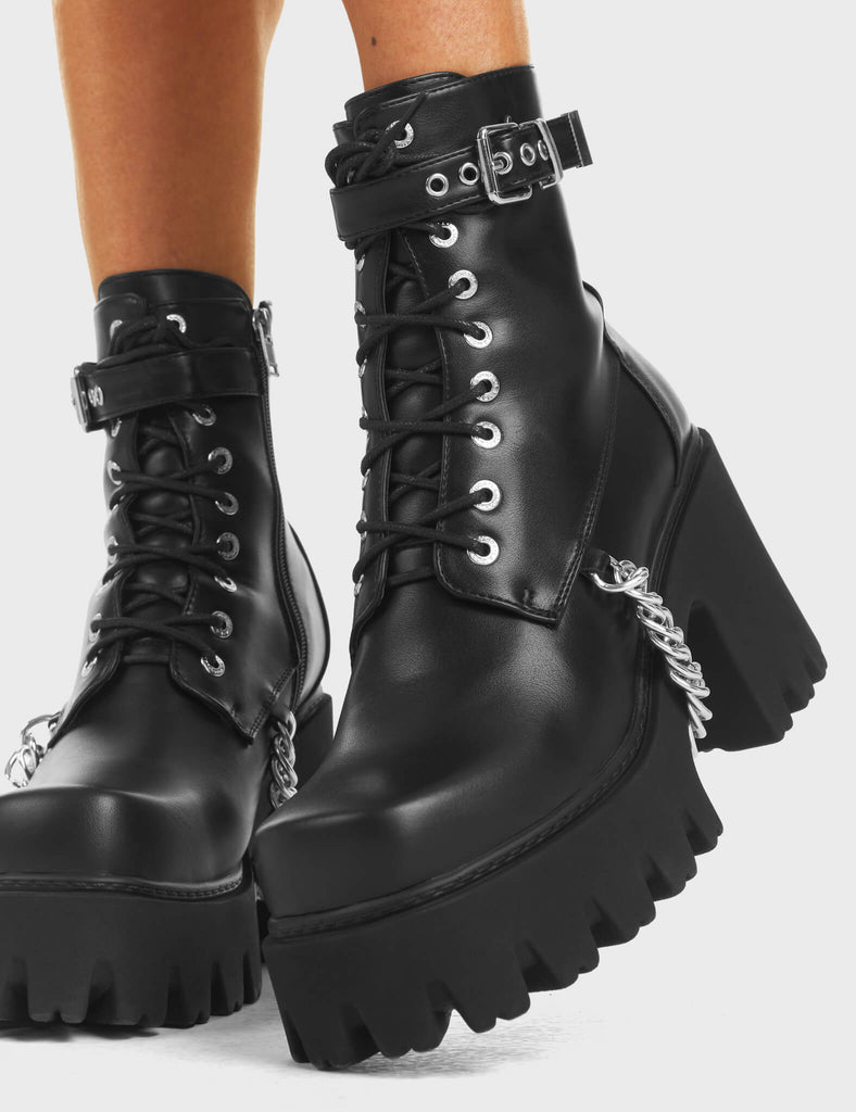 Play Hard Chunky Platform Ankle Boots in Black. Features lace-up detailing, an adjustable strap with a silver square buckle, and hanging silver chain around the sole.